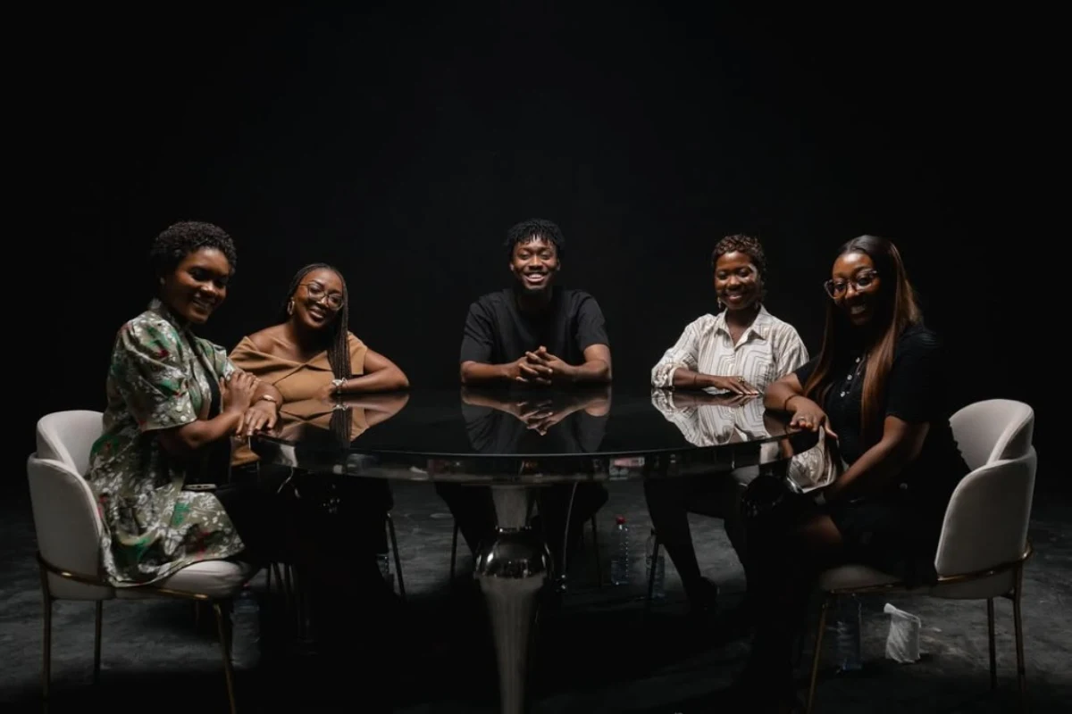 Five podcast guests seated around a round reflective table on a lit set with a dark background for an all-panel discussion.