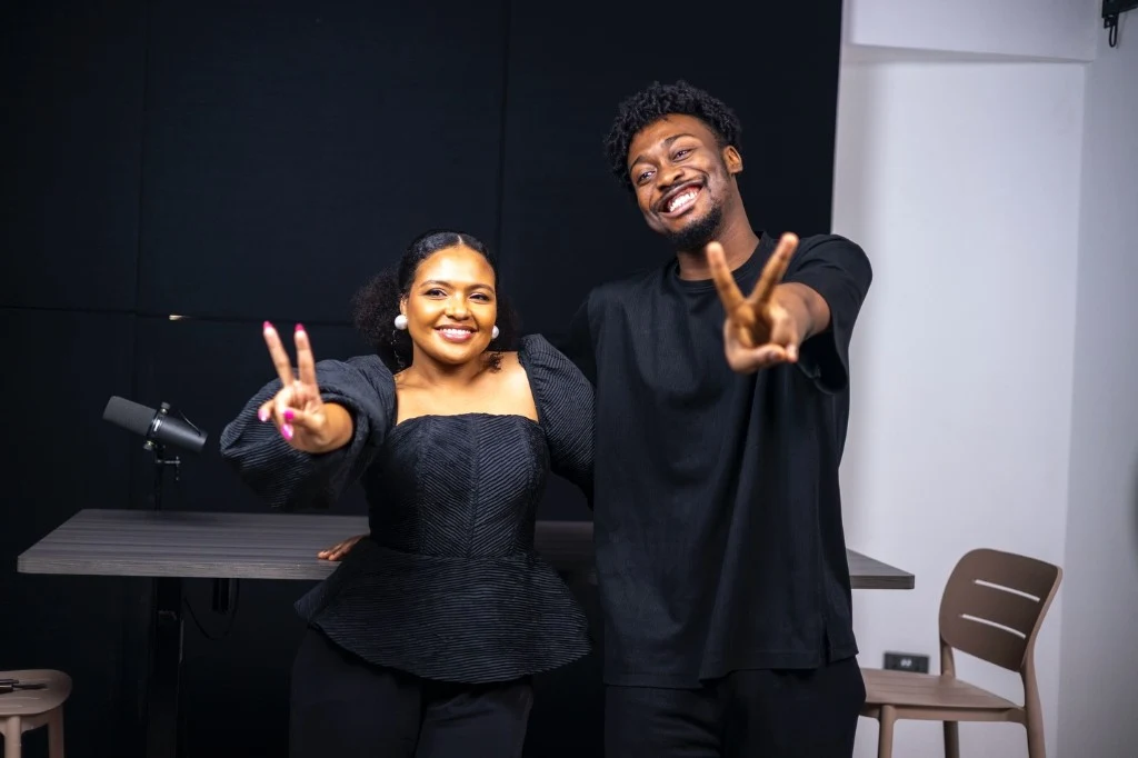 Podcast host Reagan Marfo with a guest behind the desk with a boom microphone, smiling and waving at the camera in a recording studio.