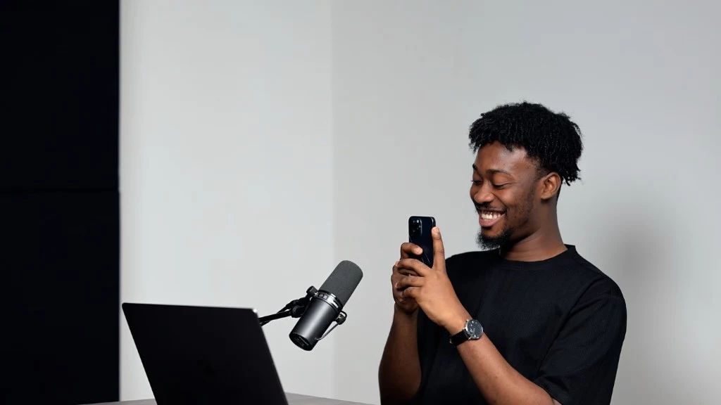 Reagan Marfo at the podcast desk with a SM7-style microphone on a boom arm and a smartphone in portrait orientation for recording short-form clips.